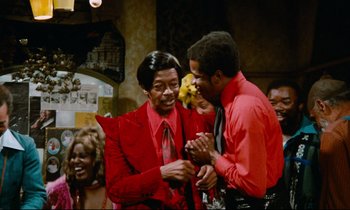 Movie still from “Don't Play Us Cheap” (1972), directed by Melvin Van Peebles – A man and a woman in red are holding hands and smiling; Medium shot, Over the shoulder angle