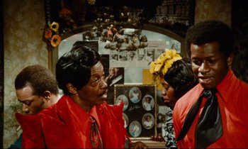 Movie still from “Don't Play Us Cheap” (1972), directed by Melvin Van Peebles – A group of people standing next to each other in a room; Medium shot, Over the shoulder angle