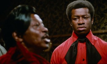 Movie still from “Don't Play Us Cheap” (1972), directed by Melvin Van Peebles – A man in a red shirt and a man in a black tie; Close Up shot, Over the shoulder angle
