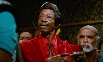 Movie still from “Don't Play Us Cheap” (1972), directed by Melvin Van Peebles – An older man wearing a red shirt and a red neck tie; Close Up shot, Low angle