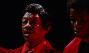 Movie still from “Don't Play Us Cheap” (1972), directed by Melvin Van Peebles – A man in a red shirt and a tie; Close Up shot, Low angle