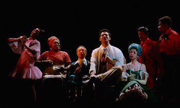 Movie still from “Don't Play Us Cheap” (1972), directed by Melvin Van Peebles – A group of men and women sitting on a stage; Wide shot, Low angle