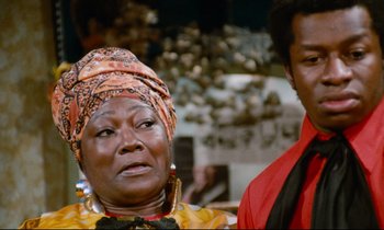 Movie still from “Don't Play Us Cheap” (1972), directed by Melvin Van Peebles – An older woman wearing a head scarf with a man standing behind her; Close Up shot, Low angle