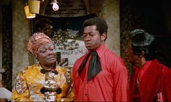 Movie still from “Don't Play Us Cheap” (1972), directed by Melvin Van Peebles – A man in a red shirt is standing next to a woman in a yellow dress; Medium shot, Over the shoulder angle