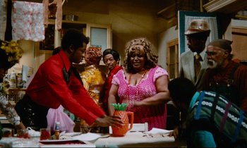 Movie still from “Don't Play Us Cheap” (1972), directed by Melvin Van Peebles – A group of people standing around a table with food; Medium shot, High angle