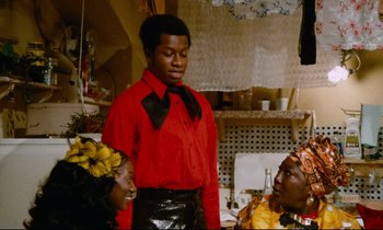 Movie still from “Don't Play Us Cheap” (1972), directed by Melvin Van Peebles – A man in a red shirt and a woman in a red shirt; Medium shot, Over the shoulder angle