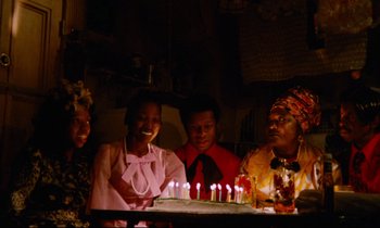 Movie still from “Don't Play Us Cheap” (1972), directed by Melvin Van Peebles – A group of people sitting around a cake with lit candles on it; Medium shot, High angle