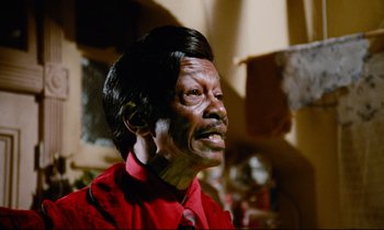 Movie still from “Don't Play Us Cheap” (1972), directed by Melvin Van Peebles – An old man with black hair and a red shirt; Close Up shot, Low angle