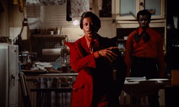 Movie still from “Don't Play Us Cheap” (1972), directed by Melvin Van Peebles – A woman in a red jacket standing in a kitchen; Medium shot, Low angle