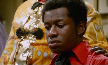 Movie still from “Don't Play Us Cheap” (1972), directed by Melvin Van Peebles – A man in a red shirt and a black tie; Close Up shot, Low angle