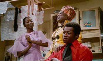 Movie still from “Don't Play Us Cheap” (1972), directed by Melvin Van Peebles – Two women and a man are in a room; Medium shot, Over the shoulder angle