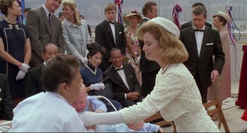 Movie still from “Double Impact” (1991), directed by Sheldon Lettich – A woman in a white dress and a hat is shaking hands with a man in a white suit; Medium shot, High angle