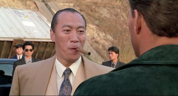 Movie still from “Double Impact” (1991), directed by Sheldon Lettich – A man smoking a cigar while another man looks on; Close Up shot, Over the shoulder angle