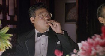 Movie still from “Double Impact” (1991), directed by Sheldon Lettich – A man in a suit and bow tie eating a piece of food; Medium shot, Low angle