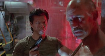 Movie still from “Double Impact” (1991), directed by Sheldon Lettich – A man holding a gun in front of another man in a red room; Close Up shot, Over the shoulder angle