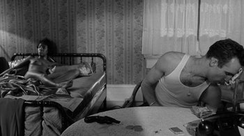 Movie still from “Down by Law” (1986), directed by Jim Jarmusch – A man sitting in front of a bed next to a table; Medium shot, High angle