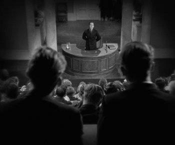 Movie still from “Dr. Jekyll and Mr. Hyde” (1931), directed by Rouben Mamoulian – A group of people sitting in front of a man in front of an audience; Wide shot, Over the shoulder angle