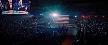 Movie still from “Don't Look Up” (2021), directed by Adam McKay – A crowd of people in a stadium with an american flag on the stage; Extreme Wide shot, High angle