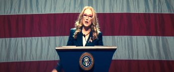 Movie still from “Don't Look Up” (2021), directed by Adam McKay – A woman is speaking at a podium in front of an american flag; Medium shot, Low angle