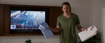 Movie still from “Draft Day” (2014), directed by Ivan Reitman – A man holding up a shirt in front of a television; Medium shot, Over the shoulder angle