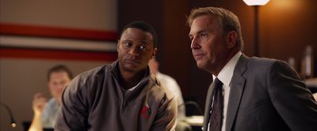 Movie still from “Draft Day” (2014), directed by Ivan Reitman – A man and a man in a suit sitting next to each other; Close Up shot, Over the shoulder angle