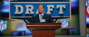 Movie still from “Draft Day” (2014), directed by Ivan Reitman – A man in a suit and tie standing at a podium; Medium shot, Low angle