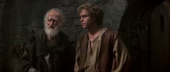 Movie still from “Dragonslayer” (1981), directed by Matthew Robbins – A man standing next to an older man in front of a stone wall; Medium shot, Over the shoulder angle