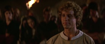 Movie still from “Dragonslayer” (1981), directed by Matthew Robbins – A man with curly blonde hair standing next to a crowd of onlookers; Close Up shot, Over the shoulder angle