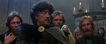 Movie still from “Dragonslayer” (1981), directed by Matthew Robbins – A man wearing a fur coat with a beard and a gold medallion; Close Up shot, Over the shoulder angle