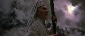 Movie still from “Dragonslayer” (1981), directed by Matthew Robbins – An old man holding a stick in front of a cloudy sky; Medium shot, Low angle