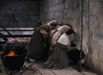 Movie still from “Drums Along the Mohawk” (1939), directed by John Ford – A group of people sitting on the ground near a fire pit; Wide shot, High angle