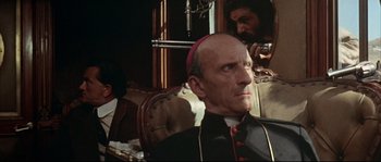 Movie still from “Duck, You Sucker!” (1971), directed by Sergio Leone – A man in a priest's outfit sitting in a chair with another man behind him; Close Up shot, Over the shoulder angle