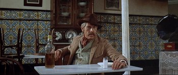 Movie still from “Duck, You Sucker!” (1971), directed by Sergio Leone – A man sitting at a table with a bottle of alcohol; Medium shot, Low angle