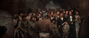Movie still from “Duck, You Sucker!” (1971), directed by Sergio Leone – A group of people in a tunnel with a man standing in front of them holding guns; Wide shot, High angle