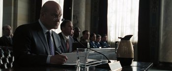 Movie still from “Eagle Eye” (2008), directed by D.J. Caruso – A group of people sitting at a table with microphones; Medium shot, Over the shoulder angle