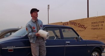 Movie still from “Eat My Dust” (1976), directed by Charles B. Griffith – A man standing next to a blue car holding a roll of toilet paper; Wide shot, Low angle