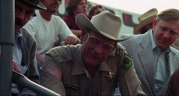 Movie still from “Eat My Dust” (1976), directed by Charles B. Griffith – An older man wearing a cowboy hat and glasses; Medium shot, Over the shoulder angle