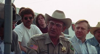 Movie still from “Eat My Dust” (1976), directed by Charles B. Griffith – An older man wearing a sheriff's hat and glasses; Medium shot, Over the shoulder angle