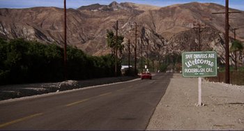 Movie still from “Eat My Dust” (1976), directed by Charles B. Griffith – A car driving down a street next to palm trees and mountains; Extreme Wide shot, High angle