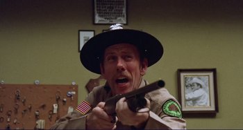 Movie still from “Eat My Dust” (1976), directed by Charles B. Griffith – A man holding a gun in a room with other people; Close Up shot, Low angle