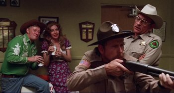 Movie still from “Eat My Dust” (1976), directed by Charles B. Griffith – A man in a uniform is smoking a cigarette while a woman in a floral dress looks on; Medium shot, Over the shoulder angle