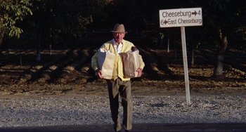 Movie still from “Eat My Dust” (1976), directed by Charles B. Griffith – An older man carrying bags of food in front of a street sign; Wide shot, Low angle