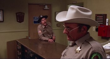 Movie still from “Eat My Dust” (1976), directed by Charles B. Griffith – A man in a sheriff's uniform sitting in front of another man; Medium shot, Over the shoulder angle