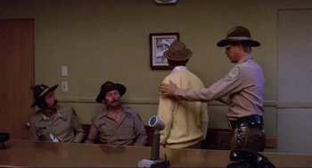 Movie still from “Eat My Dust” (1976), directed by Charles B. Griffith – A man in a hat is standing in front of a judge; Medium shot, Over the shoulder angle