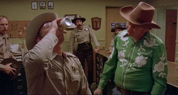 Movie still from “Eat My Dust” (1976), directed by Charles B. Griffith – A group of men standing in a room; Medium shot, Over the shoulder angle