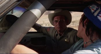 Movie still from “Eat My Dust” (1976), directed by Charles B. Griffith – A police officer sitting in the driver's seat of a vehicle; Medium shot, Over the shoulder angle