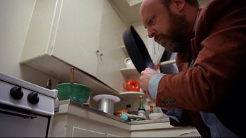 Movie still from “Eating Raoul” (1982), directed by Paul Bartel – A bearded man in an orange shirt is in the kitchen preparing a meal; Close Up shot, Low angle
