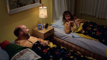 Movie still from “Eating Raoul” (1982), directed by Paul Bartel – A man and a woman laying in bed with a baby; Medium shot, High angle