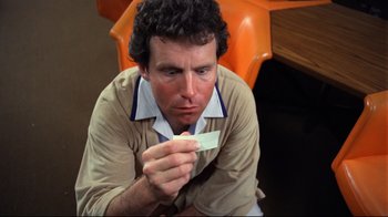 Movie still from “Eating Raoul” (1982), directed by Paul Bartel – A man sitting on the ground holding a piece of paper; Close Up shot, High angle