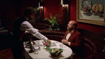 Movie still from “Eating Raoul” (1982), directed by Paul Bartel – A man sitting at a dinner table with a waiter; Wide shot, Over the shoulder angle